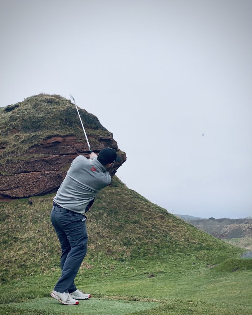 Golfer teeing off on the blind par 3 at Cullen Links, a historic course on the Old Tom Morris Trail, with rugged coastal features and dramatic elevation.