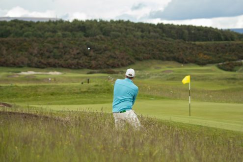Golfer playing a shot from rough beside a green at Royal Dornoch